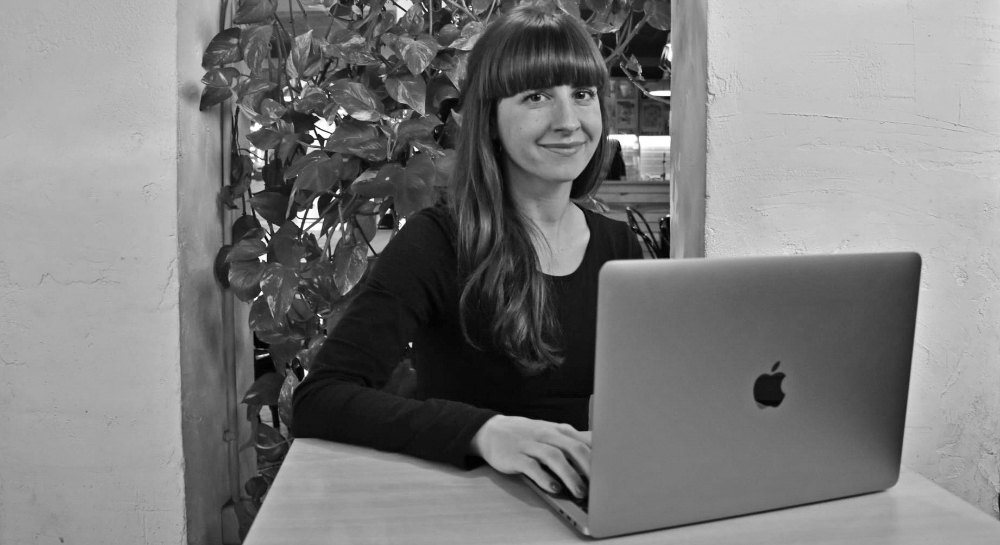 A woman weraing a black shirt, sitting at a desk, working on her Mac laptop